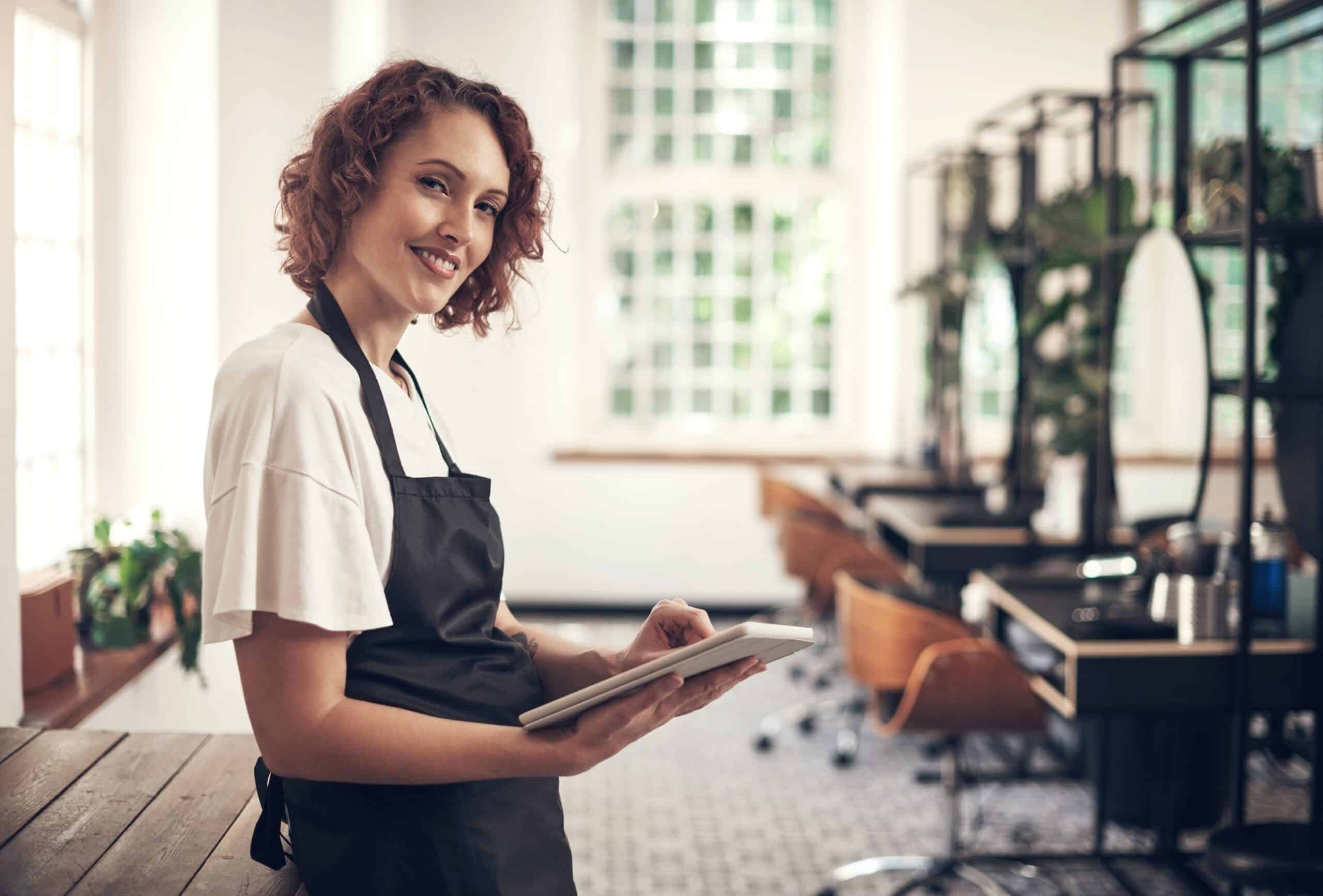 Smiling hairstylist holding tablet in modern salon.